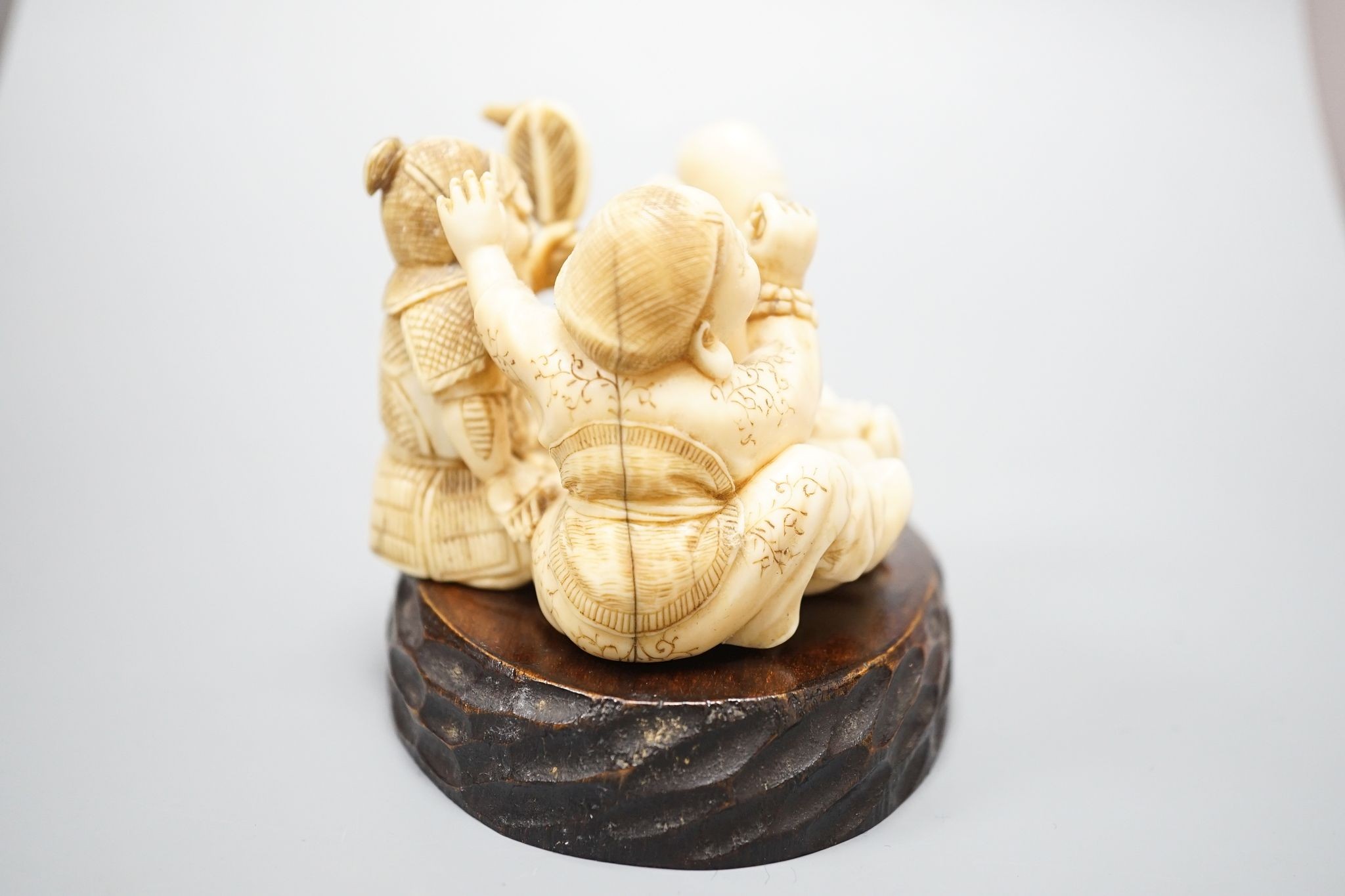 A Japanese ivory okimono of the gods Daikoku, Bishamonten and Fukurokuju, Meiji period, signed to a red lacquer tablet, 8cm wide, wood stand